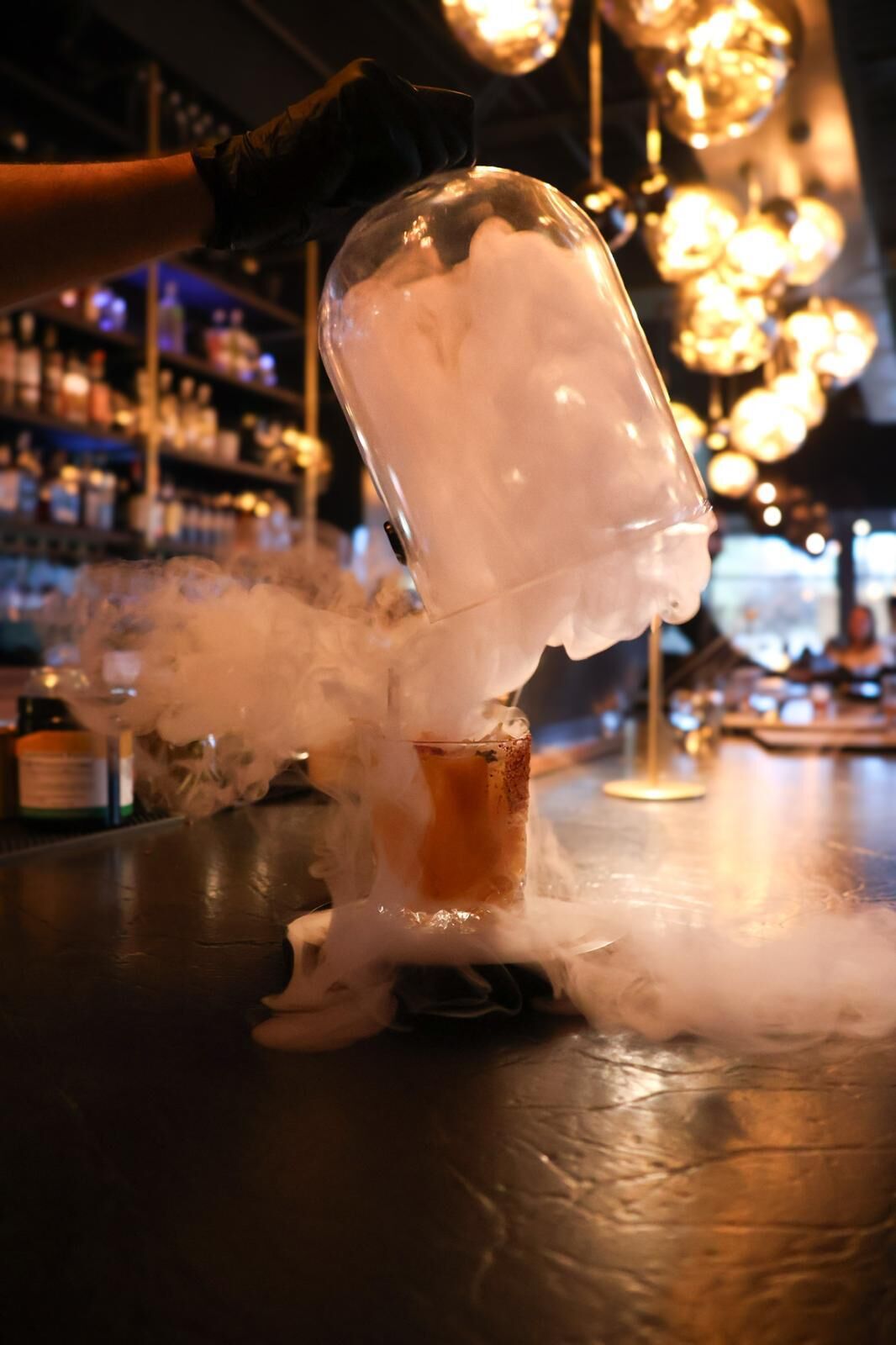 A glass cloche lifted from a cocktail, releasing a cloud of smoke across the bar